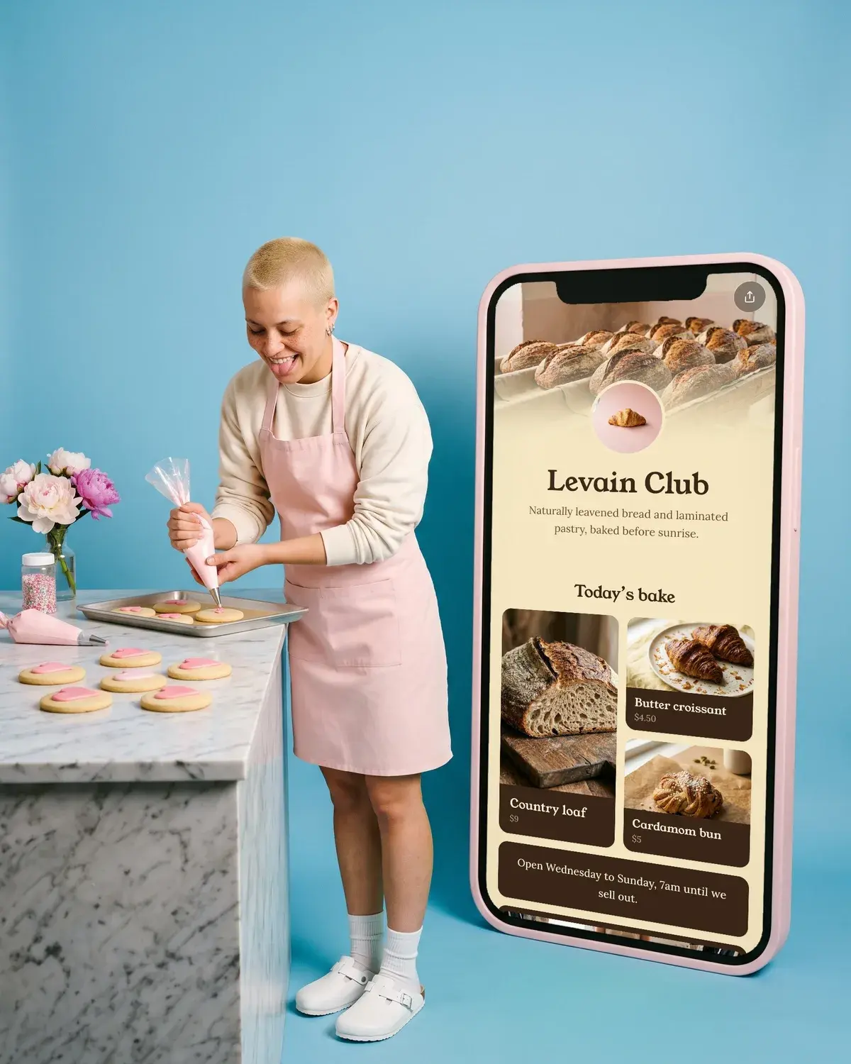 A baker pipes pink hearts onto cookies beside a giant phone showing the Levain Club page