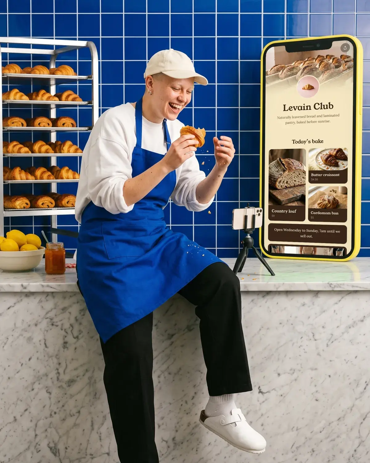 A baker films a croissant bite for TikTok beside a giant phone showing the Levain Club page