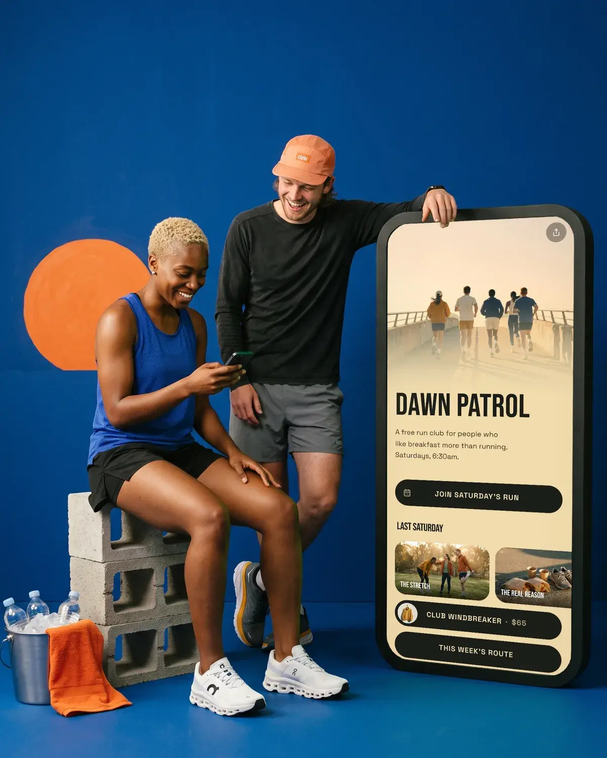 Two runners laugh on a cobalt set beside a giant phone showing the Dawn Patrol run club page