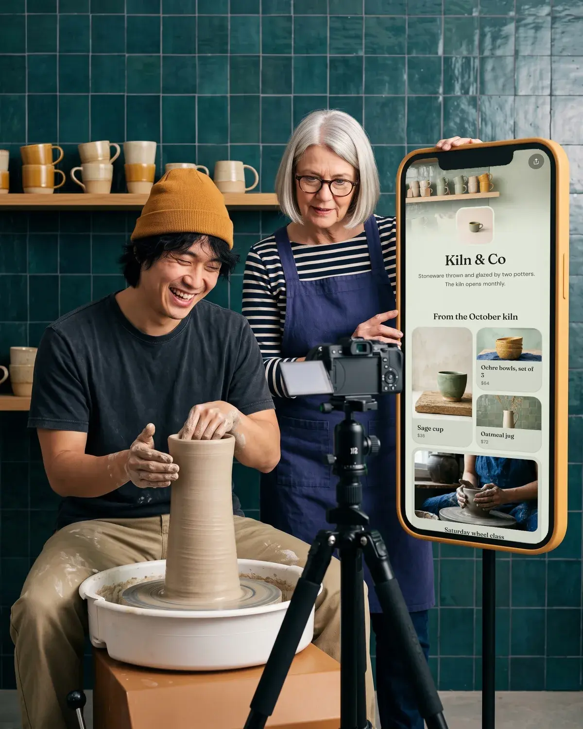 A potter throws a pot for a camera while his partner checks it, beside a giant phone showing Kiln & Co