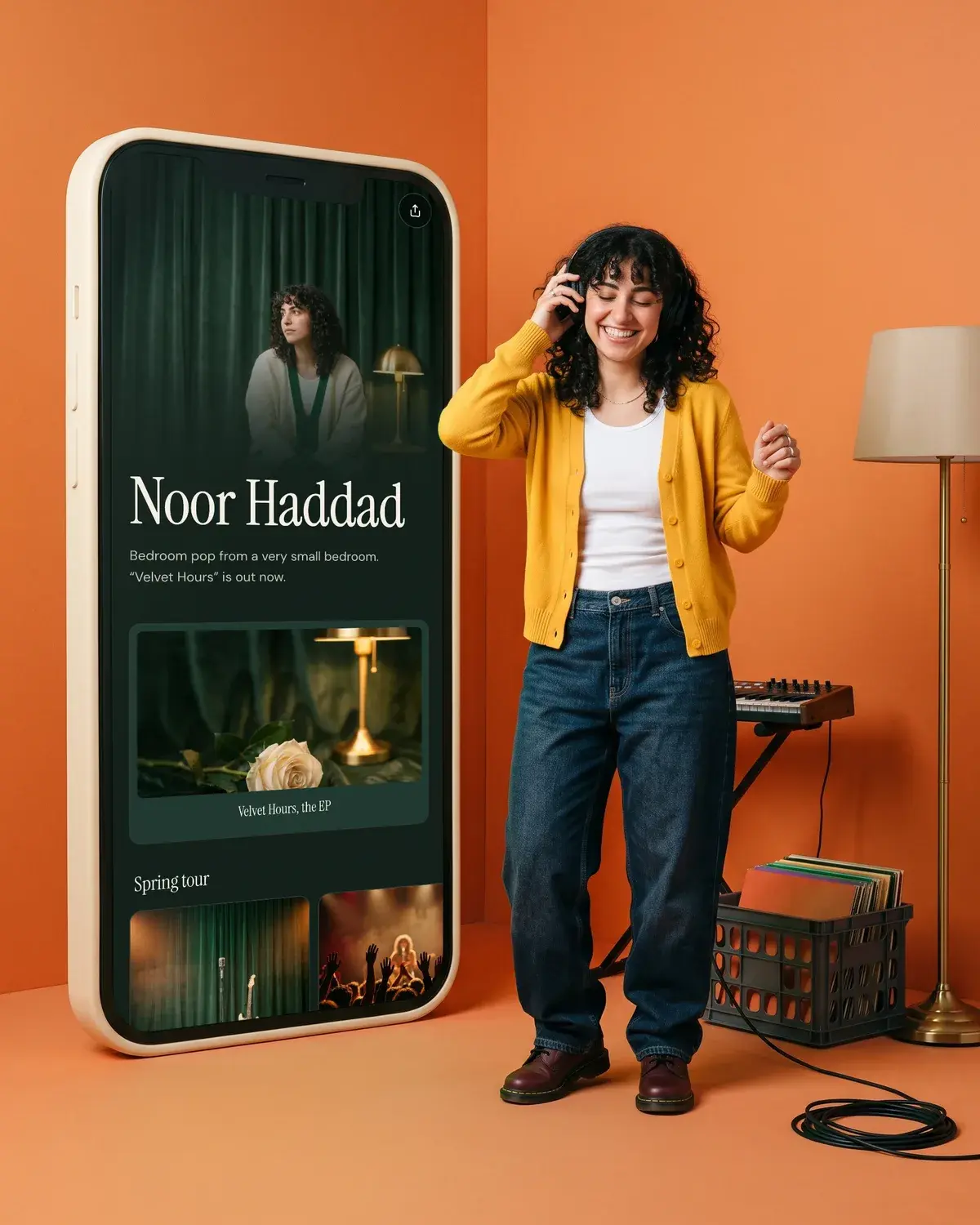 A musician dances in headphones on a tangerine set beside a giant phone showing her music page