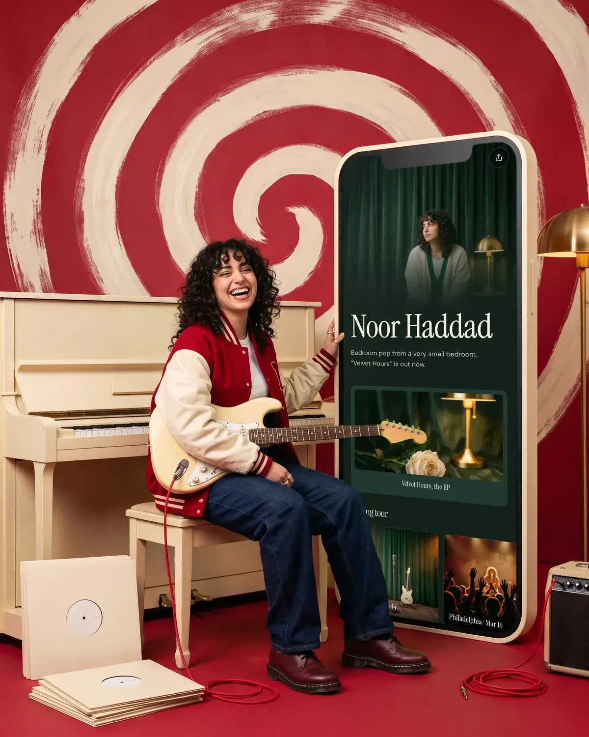 A musician laughs at a piano with a guitar beside a giant phone showing her music page