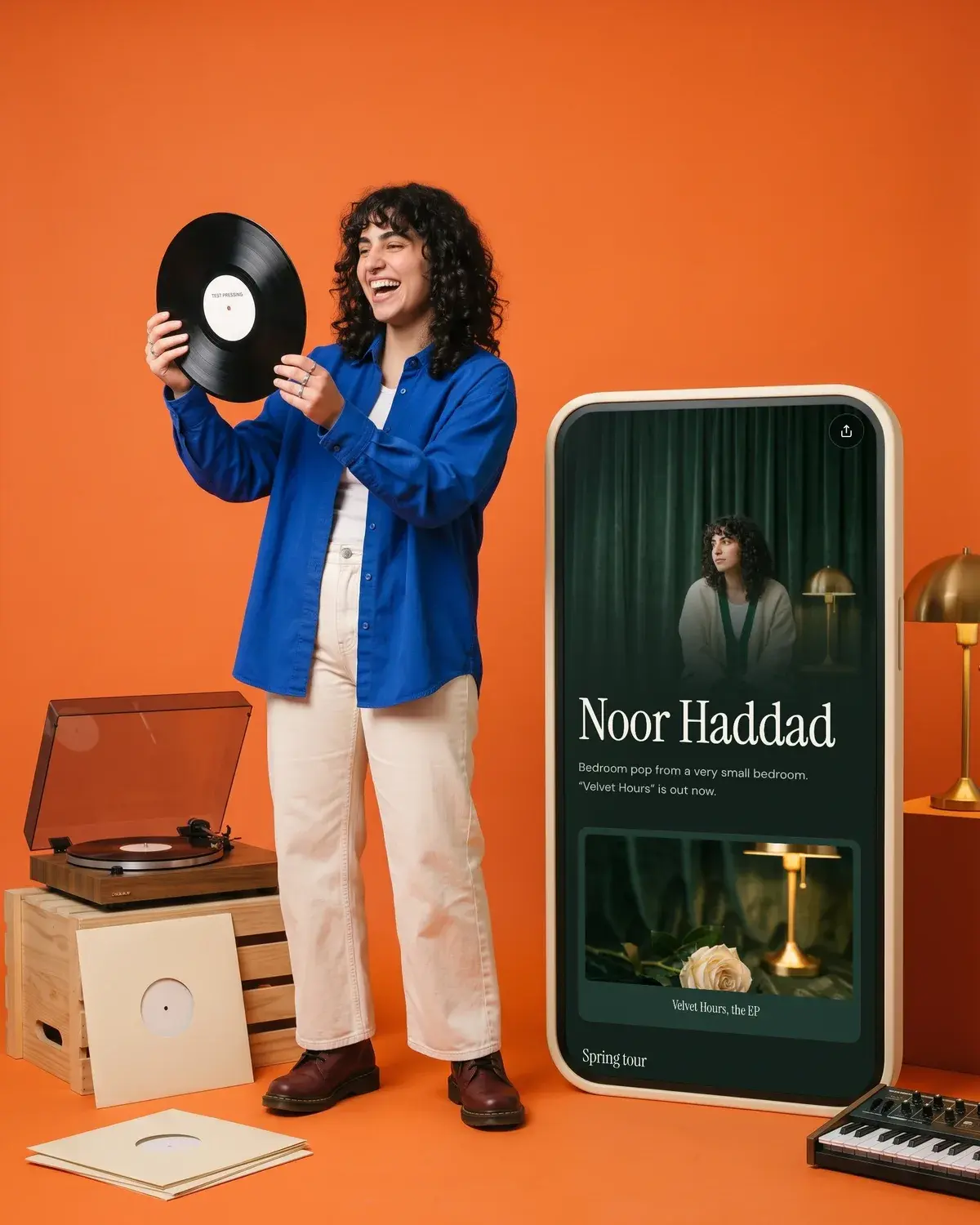 A musician holds up a vinyl record on a tangerine set beside a giant phone showing her music page