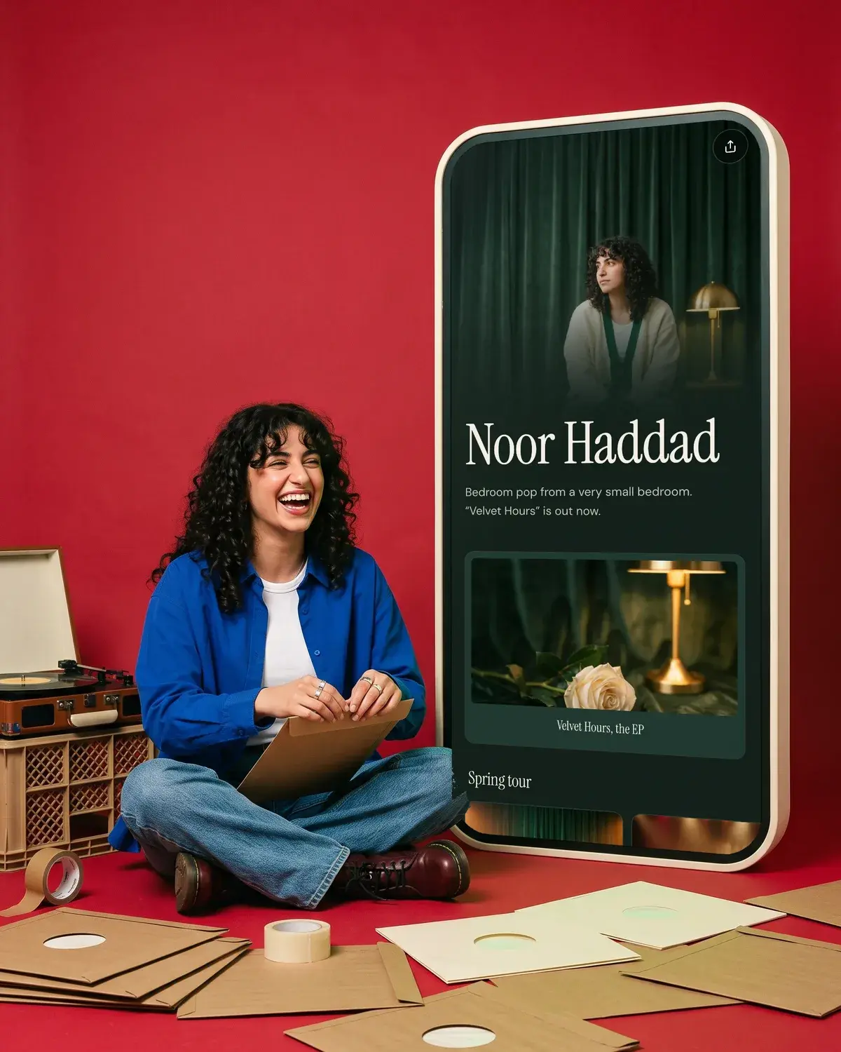 A musician seals vinyl mailers on the floor beside a giant phone showing her music page