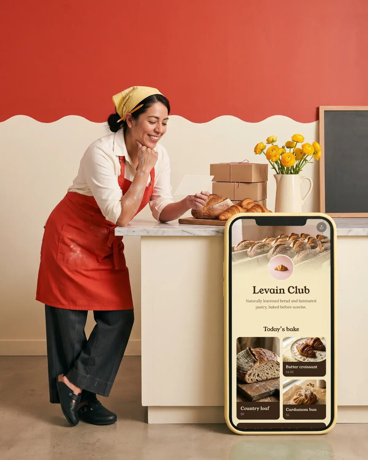 A baker leans on her counter smiling at a note beside a giant phone showing the Levain Club page