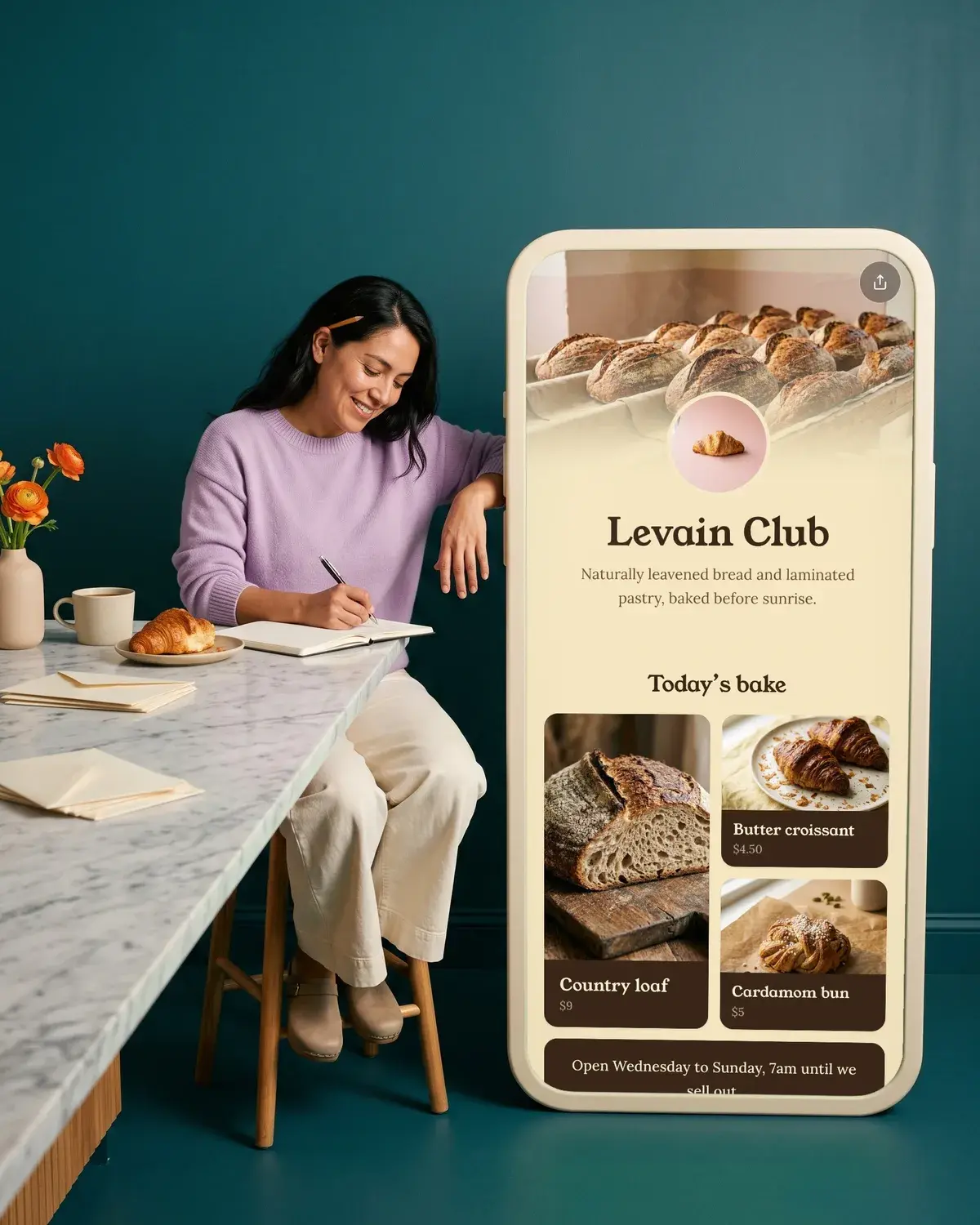 A baker writes a newsletter at the counter beside a giant phone showing the Levain Club page