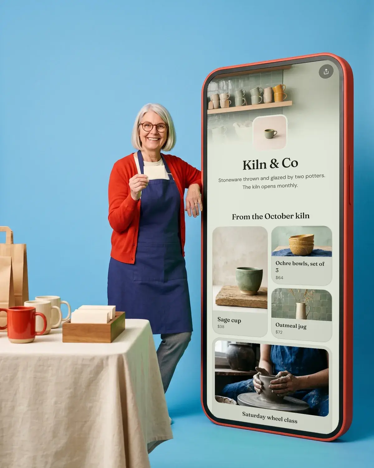 A potter holds up a business card at a craft fair beside a giant phone showing the Kiln & Co page