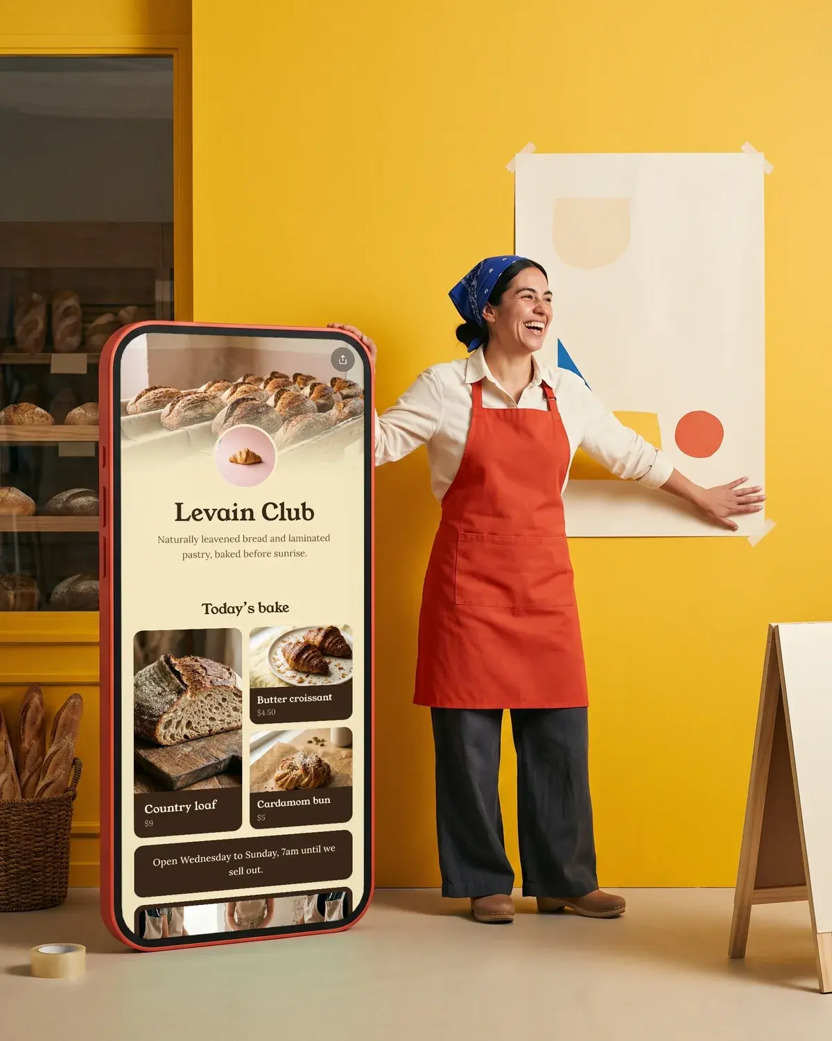 A baker tapes a poster to the bakery window beside a giant phone showing the Levain Club page