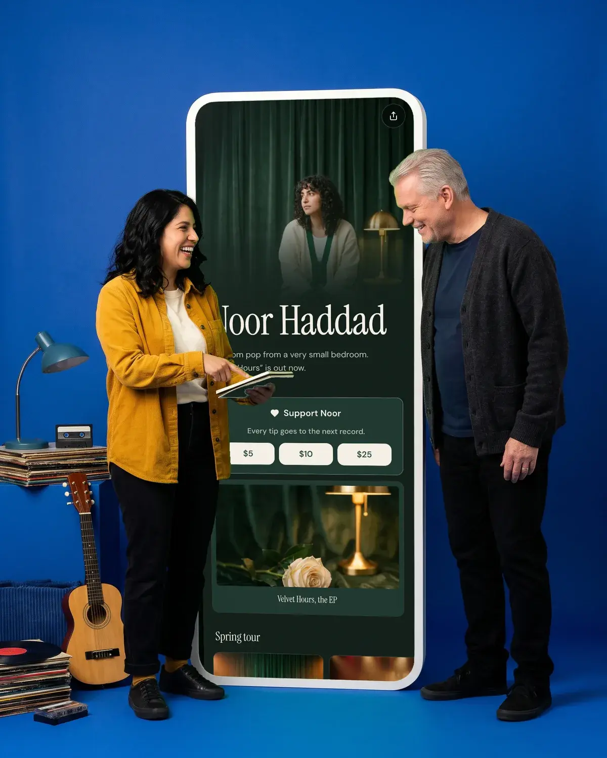 Two musicians laugh over a release notebook beside a giant phone prop against a cobalt blue backdrop
