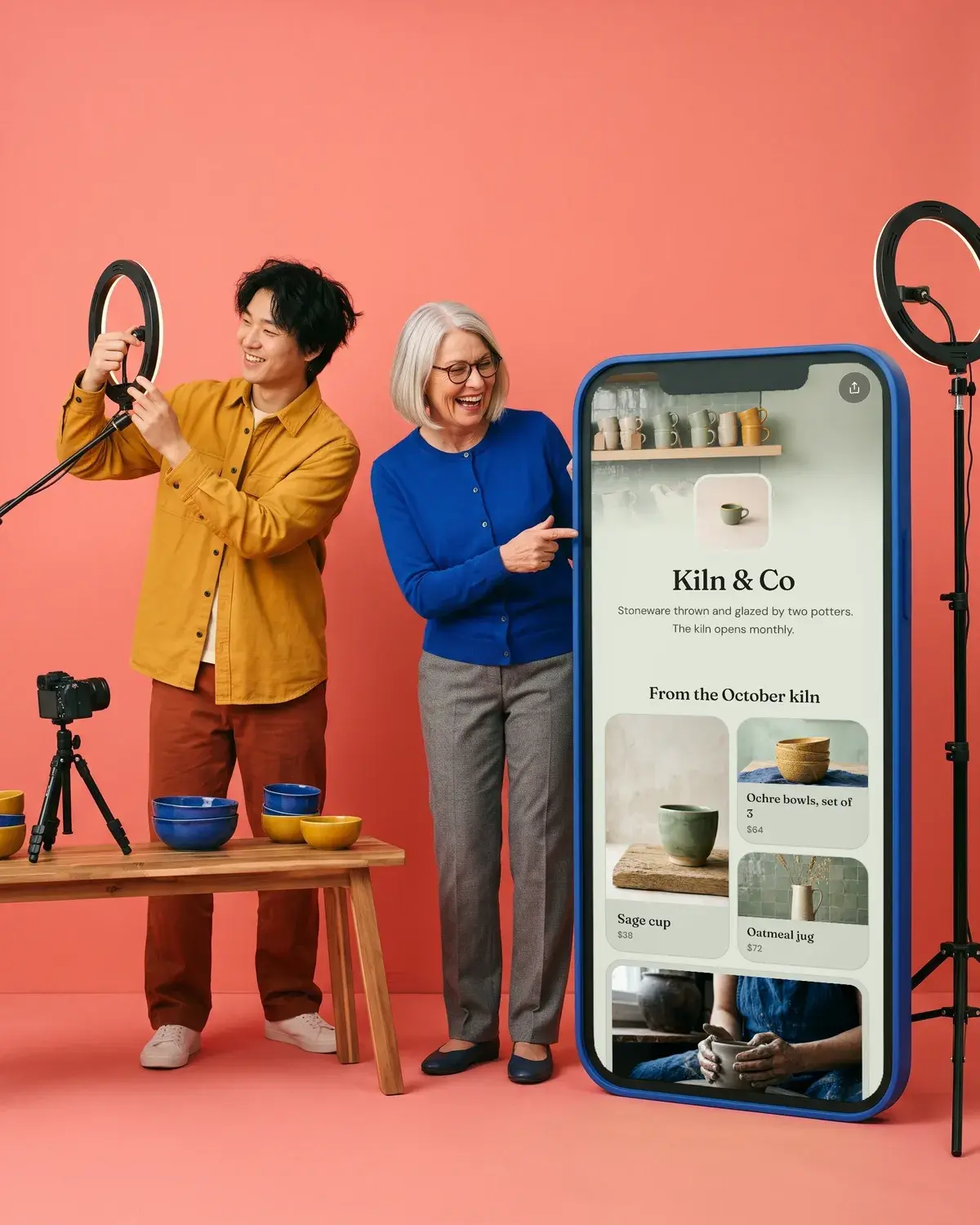 Two potters set up a ring light beside a giant phone showing the Kiln & Co page